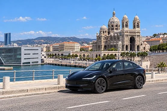 Tesla noire stationnée près d’un plan d’eau, devant un grand bâtiment à deux tours.
