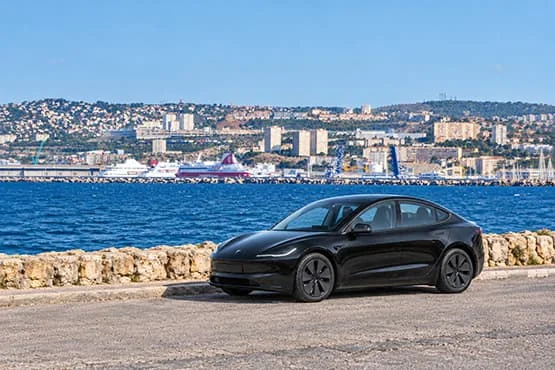 Tesla noire au bord de la mer, devant un panorama de ville et de collines.