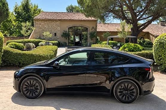 Tesla noire de profil devant une maison en pierre et un jardin.